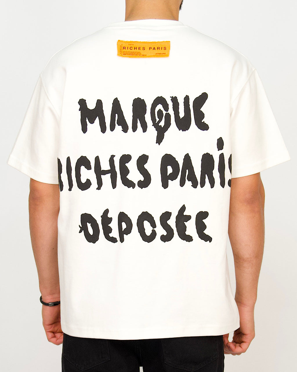 Back view of a men’s oversized white T-shirt featuring bold black Riches Paris typography and a contrast neck patch.