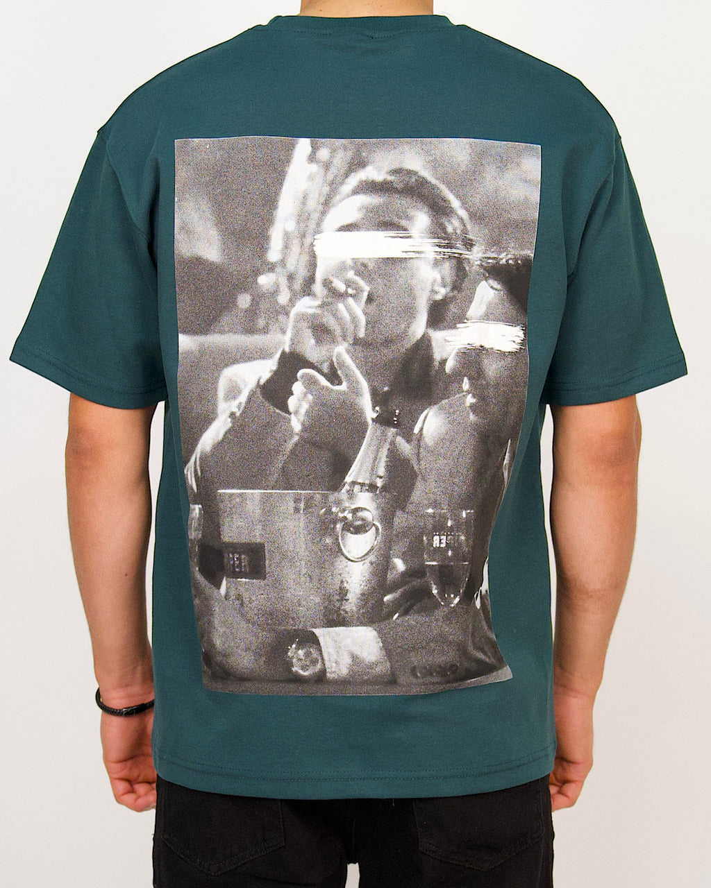 Back view of a men’s oversized green T-shirt with a large monochrome photo-print featuring blurred faces.