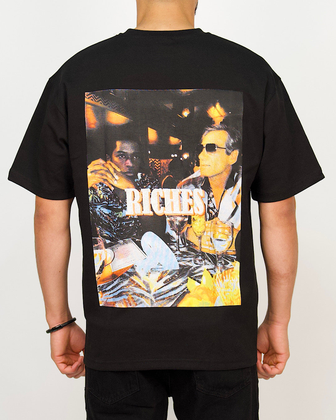 Back view of a men’s oversized black T-shirt featuring a colourful nightlife photo-print with “RICHES” branding.