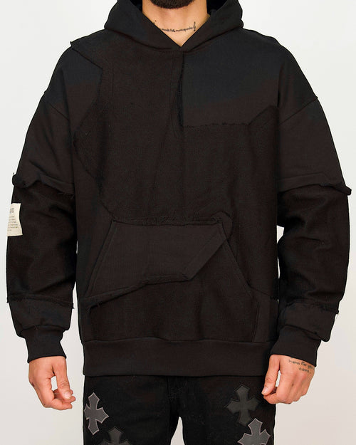 Front view of a men’s oversized black hoodie featuring raw-edged multi-panel construction and a sculpted kangaroo pocket.